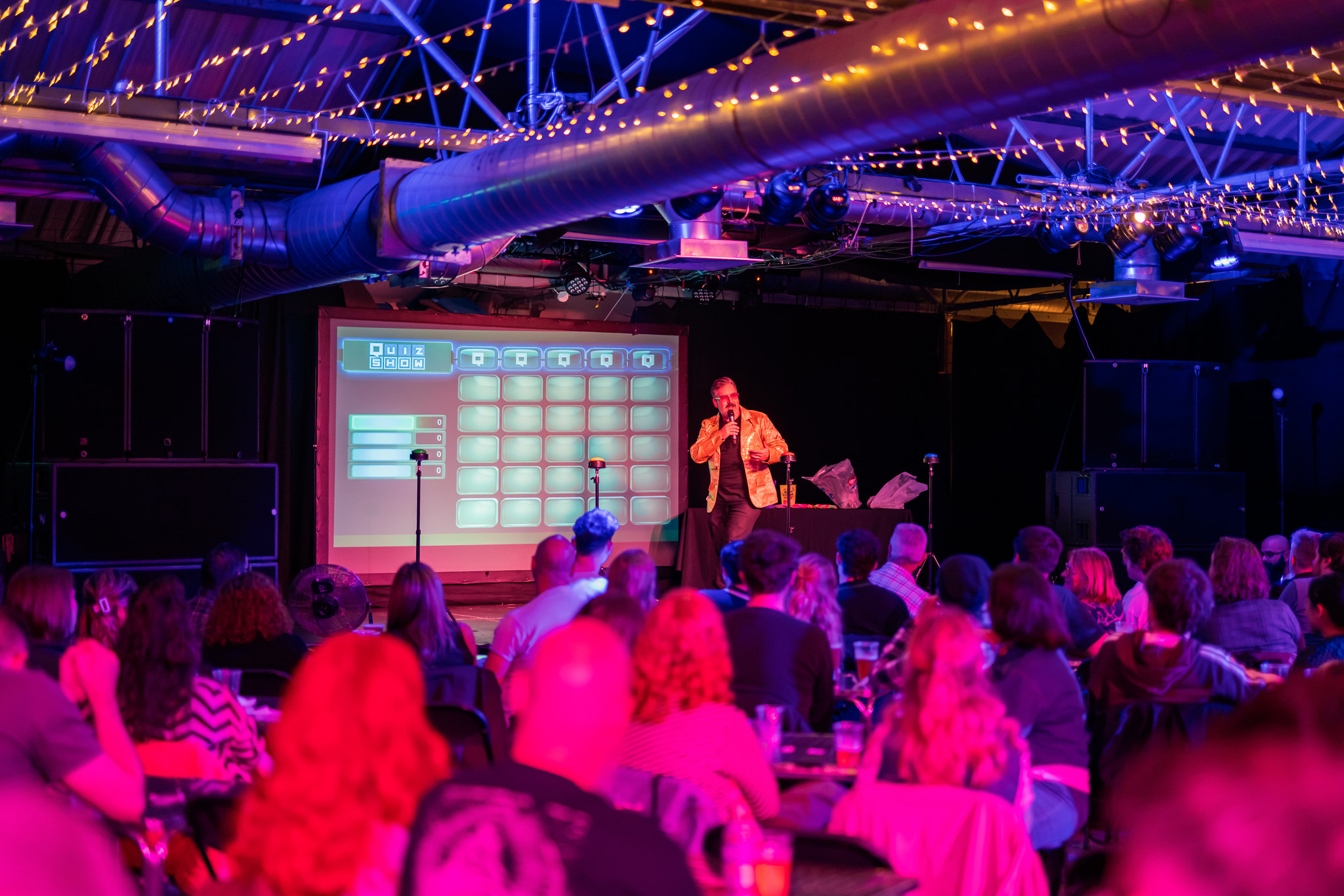 Gary on stage beside a giant quiz board, hosting a packed room under fairy lights